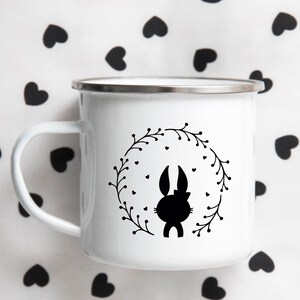 May include: White enamel mug with a black silhouette of a bunny rabbit inside a wreath. The wreath is made of black leaves and has small black hearts around it.