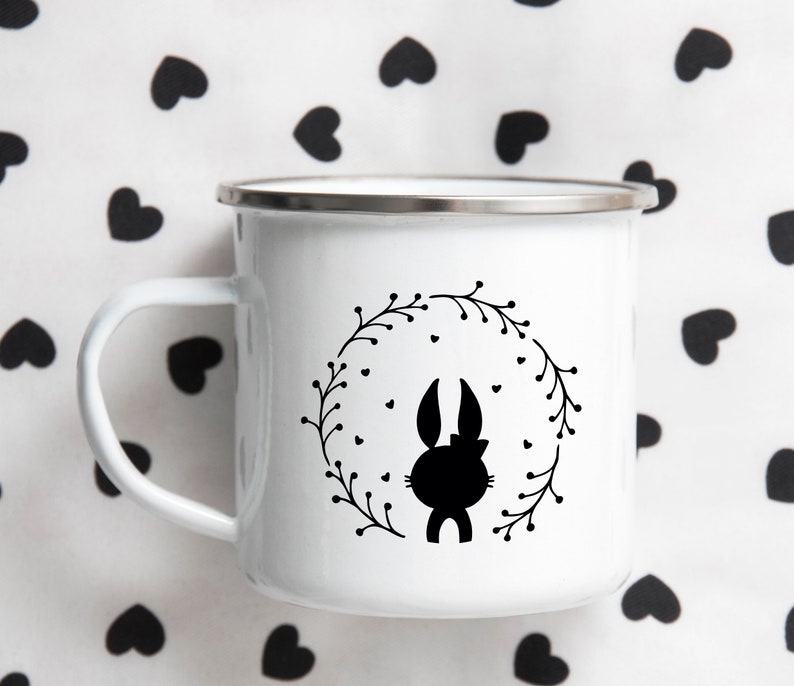 May include: White enamel mug with a black silhouette of a bunny rabbit inside a wreath. The wreath is made of black leaves and has small black hearts around it.