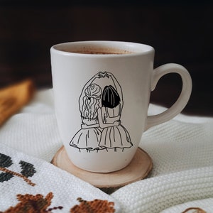 K&ouml;nnte beinhalten: Wei&szlig;e Keramiktasse mit einer schwarzen Linienzeichnung von zwei Frauen, die sich an den H&auml;nden halten und einander gegen&uuml;berstehen. Die Frauen tragen Kleider und haben langes Haar.