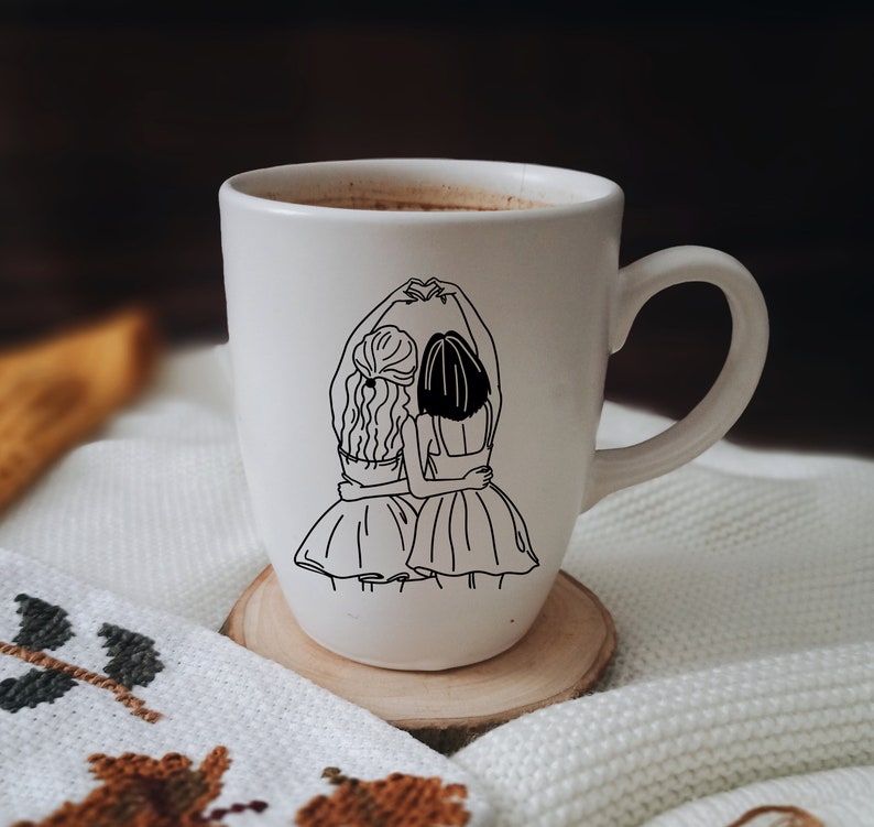 K&ouml;nnte beinhalten: Wei&szlig;e Keramiktasse mit einer schwarzen Linienzeichnung von zwei Frauen, die sich an den H&auml;nden halten und einander gegen&uuml;berstehen. Die Frauen tragen Kleider und haben langes Haar.