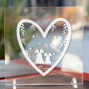 May include: A white heart-shaped design on a clear acrylic plaque with two bunnies inside the heart. The bunnies are facing each other and have small, simple features. The heart is decorated with small white stars and floral designs.