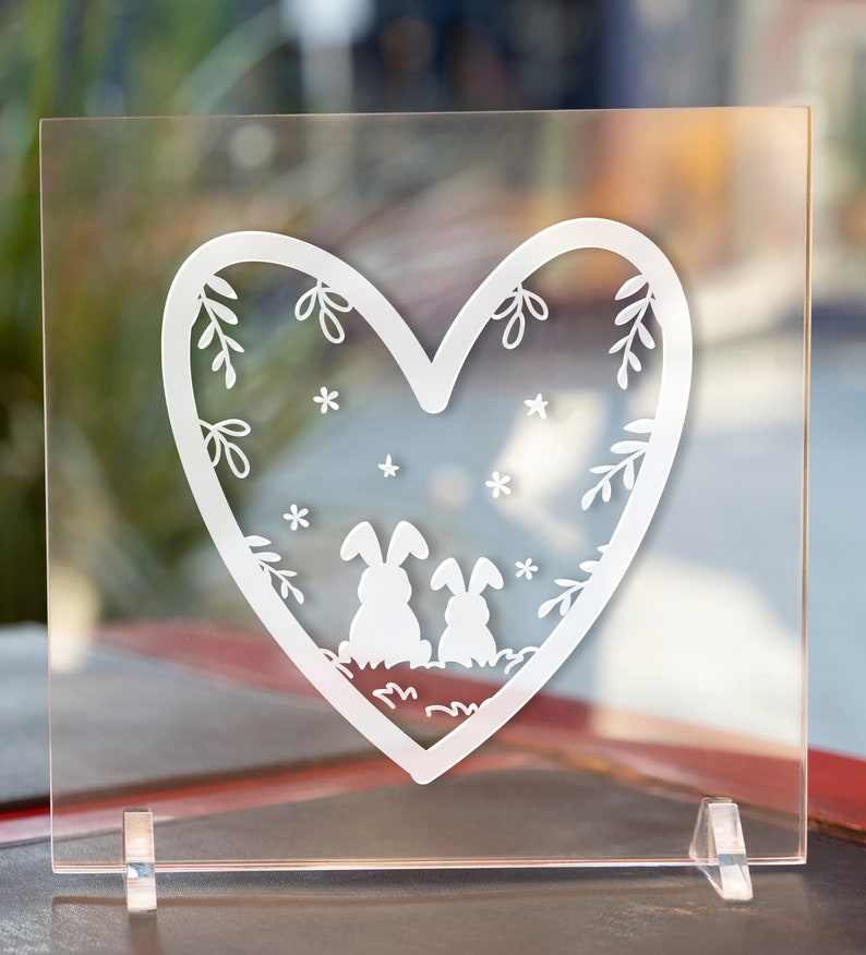May include: A white heart-shaped design on a clear acrylic plaque with two bunnies inside the heart. The bunnies are facing each other and have small, simple features. The heart is decorated with small white stars and floral designs.