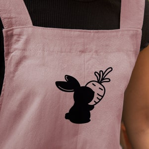May include: A pink cotton apron with adjustable straps features a black silhouette of a rabbit holding a carrot.
