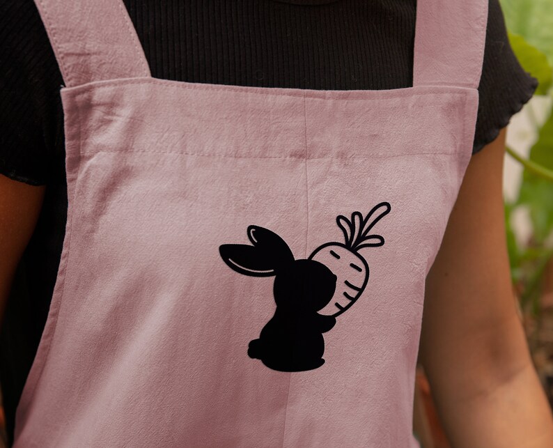 May include: A pink cotton apron with adjustable straps features a black silhouette of a rabbit holding a carrot.