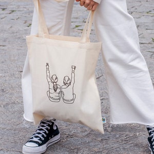 K&ouml;nnte beinhalten: Eine wei&szlig;e Canvas-Tragetasche mit zwei Figuren in schwarzer Linienkunst, die ihre Arme in die Luft heben. Die Figuren tragen einfache Kleidung und haben dem Betrachter den R&uuml;cken zugewandt. Die Tasche hat einen schwarzen Griff und ein kleines gr&uuml;nes Etikett in der unteren rechten Ecke.