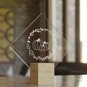 Puede incluir: Una placa de vidrio transparente en forma de diamante con un dise&ntilde;o grabado en blanco de calabazas y follaje de oto&ntilde;o. La placa est&aacute; sobre una base de madera.
