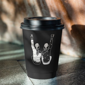 K&ouml;nnte beinhalten: Schwarzer Coffee-to-go-Becher mit einer wei&szlig;en Grafik von zwei Personen, die die Arme in die Luft strecken. Die Grafik zeigt zwei Personen mit dem R&uuml;cken zum Betrachter.