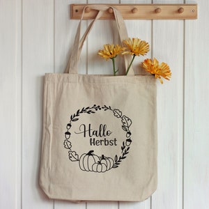 Puede incluir: Una bolsa de lona beige con asas negras. La bolsa tiene un dise&ntilde;o gr&aacute;fico negro de una corona con hojas, bellotas y calabazas. El texto "Hallo Herbst" est&aacute; dentro de la corona.