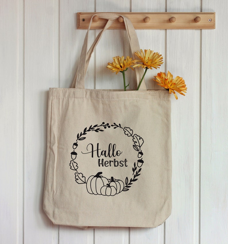 Puede incluir: Una bolsa de lona beige con asas negras. La bolsa tiene un dise&ntilde;o gr&aacute;fico negro de una corona con hojas, bellotas y calabazas. El texto "Hallo Herbst" est&aacute; dentro de la corona.