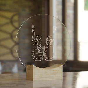 K&ouml;nnte beinhalten: Ein klarer Glaskreis mit einem Holzsockel, der ein lasergraviertes Design von zwei Figuren mit erhobenen Armen zeigt. Die Figuren sind vom Betrachter abgewandt und deuten auf Freundschaft oder Feierlichkeit hin.