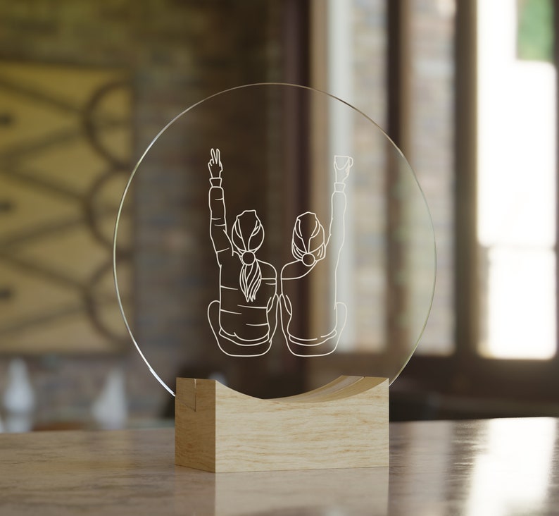K&ouml;nnte beinhalten: Ein klarer Glaskreis mit einem Holzsockel, der ein lasergraviertes Design von zwei Figuren mit erhobenen Armen zeigt. Die Figuren sind vom Betrachter abgewandt und deuten auf Freundschaft oder Feierlichkeit hin.