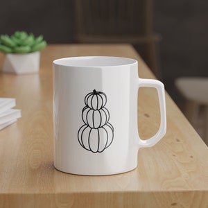 Puede incluir: Taza de cer&aacute;mica blanca con un contorno negro de tres calabazas apiladas. La taza tiene un asa en el lado derecho.