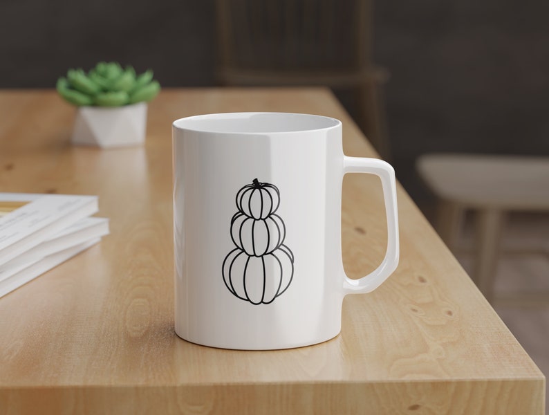 Puede incluir: Taza de cer&aacute;mica blanca con un contorno negro de tres calabazas apiladas. La taza tiene un asa en el lado derecho.
