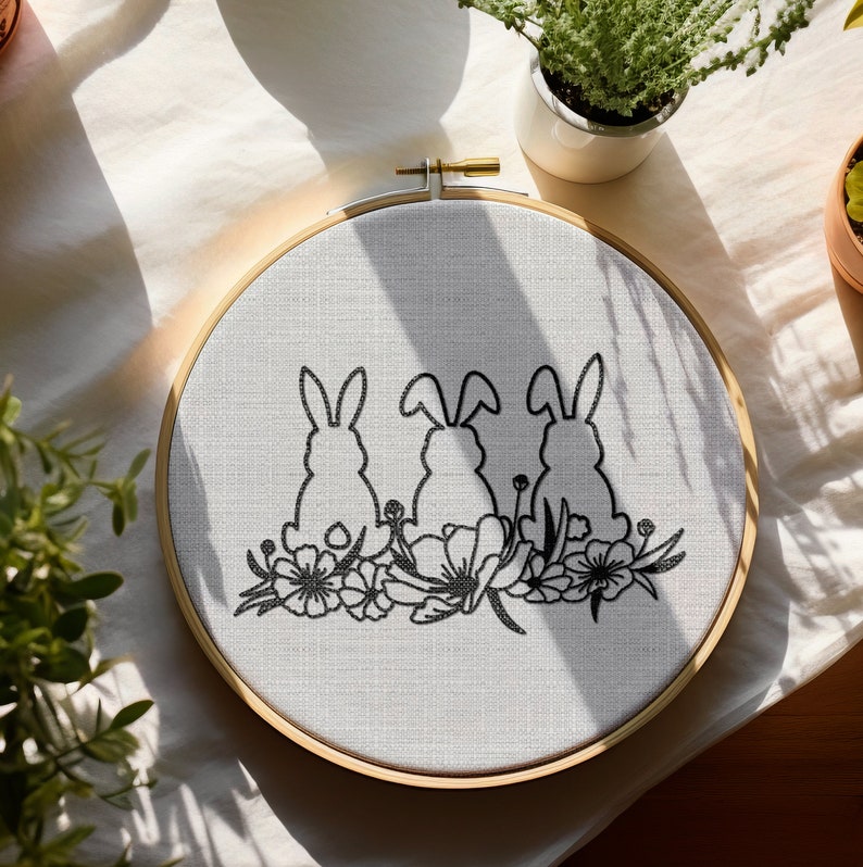 May include: A white embroidery hoop with a black embroidered design of three bunnies sitting in a row with flowers behind them.
