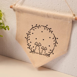May include: A beige canvas wall hanging with a black outline of two bunnies sitting inside a wreath of leaves and hearts.