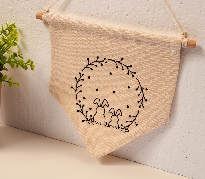 May include: A beige canvas wall hanging with a black outline of two bunnies sitting inside a wreath of leaves and hearts.