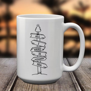 Puede incluir: Taza de cer&aacute;mica blanca con un gr&aacute;fico negro de una gaviota sobre un poste de se&ntilde;alizaci&oacute;n. El poste dice "ALL I NEED IS VITAMIN SEA" en letras may&uacute;sculas negras. La taza tiene un asa grande y est&aacute; sobre una superficie de madera.