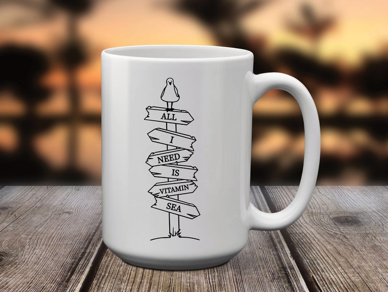 Puede incluir: Taza de cer&aacute;mica blanca con un gr&aacute;fico negro de una gaviota sobre un poste de se&ntilde;alizaci&oacute;n. El poste dice "ALL I NEED IS VITAMIN SEA" en letras may&uacute;sculas negras. La taza tiene un asa grande y est&aacute; sobre una superficie de madera.
