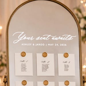 Könnte beinhalten: Ein goldgerahmter Sitzplan mit dem Schriftzug "Your seat awaits" in geschwungener Schrift. Darunter steht "ASHLEY & JAROD | MAY 24, 2026." Weiße Karten sind mit goldenen Wachssiegeln befestigt, die Tischnummern anzeigen.