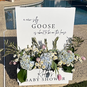 Puede incluir: Un letrero blanco con el texto "a new silly GOOSE is about to be on the loose" y "Khloe's BABY SHOWER". El letrero está decorado con hortensias azules, flores blancas y tulipanes rosas.