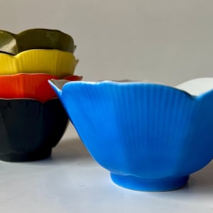 Puede incluir: Un conjunto de cuencos con forma de flor de varios colores. Los cuencos están apilados, con colores como el negro, el naranja, el amarillo y el verde. Un solo cuenco azul está colocado por separado, con el interior blanco.