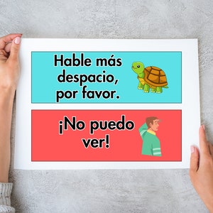 May include: Two colourful signs with Spanish text. The top sign is blue and says "Hable m&aacute;s despacio, por favor." with a cartoon tortoise. The bottom sign is red and says "&iexcl;No puedo ver!" with a cartoon person looking over their shoulder.