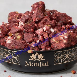 May include: A black ceramic bowl with a gold lotus flower graphic and the text MonJad, filled with a mound of irregular, dark red, textured, and crumbly pieces of freeze-dried fruit. The bowl features a repeating geometric pattern in gold.