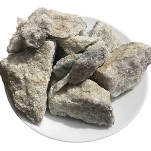 Può includere: Una collezione di rocce ruvide, grigio chiaro, con sottili sfumature viola e marroni, presentate su un piatto bianco. Le rocce hanno una superficie strutturata e dimensioni variabili, suggerendo un'estetica naturale e non raffinata.