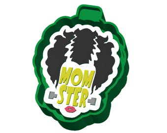 May include: A green plastic cookie cutter shaped like a head with black hair, featuring the text MOM STER in yellow letters, a pair of small grey bolts on the sides, and pink lips at the bottom, all set against a white background.