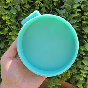 May include: A round, light teal-colored silicone mold with a small handle. The mold is held in a hand, with a background of green foliage.