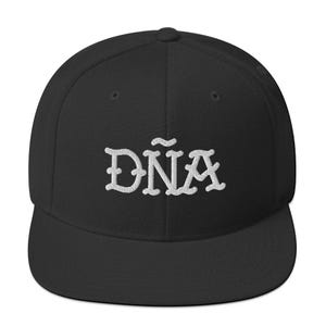 Op de afbeelding: Zwarte baseballcap met het woord "DÑA" in het wit geborduurd. De cap heeft een platte klep en een gestructureerde pasvorm. De tekst is in een decoratief, gestileerd lettertype.