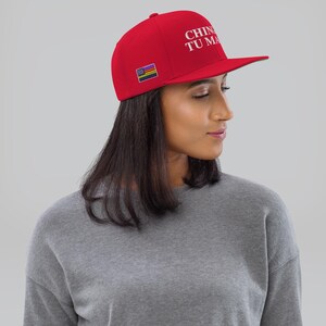 K&ouml;nnte beinhalten: Eine rote Baseballkappe mit einer wei&szlig;en Stickerei, die "CHINESE TU MA" lautet, und einem kleinen Regenbogen-Pride-Flaggen-Patch. Die Kappe wird von einer Person mit langen dunklen Haaren getragen.