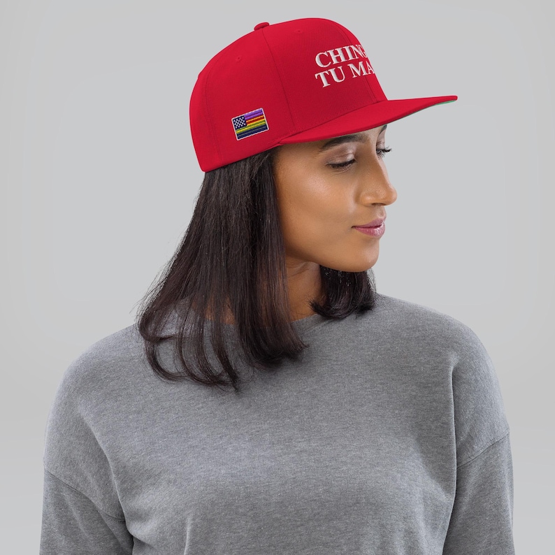 K&ouml;nnte beinhalten: Eine rote Baseballkappe mit einer wei&szlig;en Stickerei, die "CHINESE TU MA" lautet, und einem kleinen Regenbogen-Pride-Flaggen-Patch. Die Kappe wird von einer Person mit langen dunklen Haaren getragen.