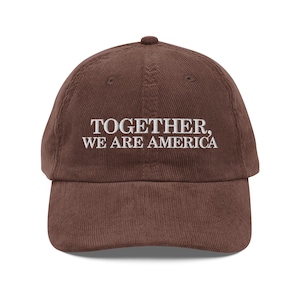 May include: A brown corduroy baseball cap with the embroidered text "TOGETHER, WE ARE AMERICA" in white. The cap features a curved peak and a small button on top. This hat is a stylish accessory.