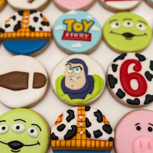 Può includere: Una collezione di biscotti di zucchero decorati con personaggi e temi del film d'animazione "Toy Story". I biscotti sono rotondi con vari disegni, tra cui Woody, Buzz Lightyear e il logo di Toy Story. I colori includono blu, giallo, rosso e verde.