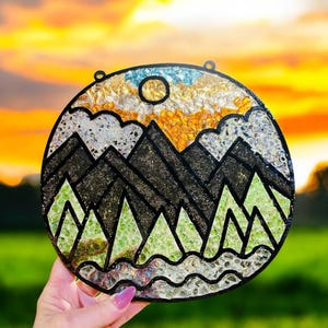 May include: A circular stained glass artwork depicting a mountain landscape. The design features black mountains, green trees, and a sky of blue, gold, and white. A yellow sun is visible. It has two small loops for hanging, and is approximately 15 cm in diameter.