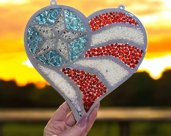 May include: Heart-shaped wall hanging with a patriotic design. The heart features a star made of blue glass and stripes of red and white glass, resembling the American flag. The metal frame has a small loop for hanging.