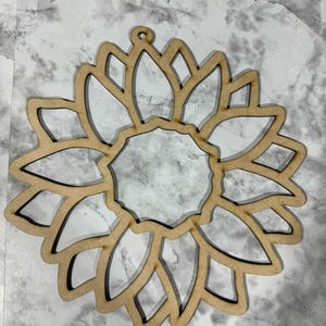 May include: A wooden sunflower cutout with a central circular opening and petal-shaped cutouts. The sunflower has a small hole at the top for hanging. The cutout is made of light-colored wood.