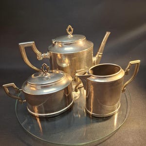 Könnte beinhalten: Ein silbernes Teeservice mit Teekanne, Milchkännchen und Zuckerdose. Das Set befindet sich auf einem durchsichtigen Glastablett.