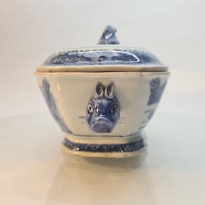 Può includere: Una ciotola con coperchio in ceramica bianca e blu con una testa di coniglio decorativa sul davanti. La ciotola presenta disegni dipinti di blu e una piccola maniglia sul coperchio. La ciotola poggia su una piccola base.