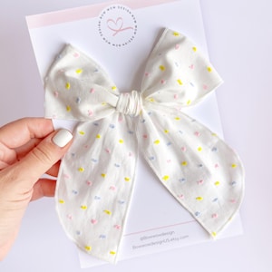 May include: A white fabric bow with a polka dot pattern in pink, yellow, and blue. The bow is tied with a white ribbon and has a slightly frayed edge.