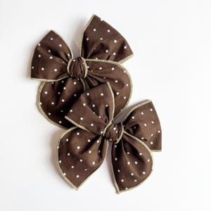 May include: Three brown fabric bows with white polka dots are arranged on a white background. Each bow has a contrasting light brown trim. The bows are tied in the center with a matching fabric knot, creating a classic, handmade look.