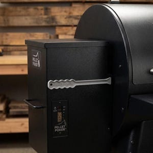 May include: A black pellet grill with a hopper and control panel. The hopper has the text "PELLET POKER" and a handle. The control panel displays a temperature gauge and the text "PELLET POKER". A gray poker tool is attached to the side.
