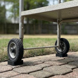 May include: A close-up of a metal utility cart with two black tires. The tires are mounted on a silver axle and sit on black rectangular blocks. The cart has a gray shelf and a silver frame. The background is a brick patio and green grass.