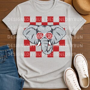 May include: A light gray t-shirt featuring a cartoon elephant wearing sunglasses with the text "Roll Tide." The elephant is set against a red and white checkered background. The words "Designed by Bun" are printed around the top of the shirt.