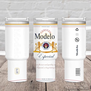 Op de afbeelding: Witte geïsoleerde beker met een handvat, met het Modelo bierlogo en tekst. De beker heeft een inhoud van ongeveer 1,18 liter. Het ontwerp bevat rode en gouden accenten en een recyclingsymbool.