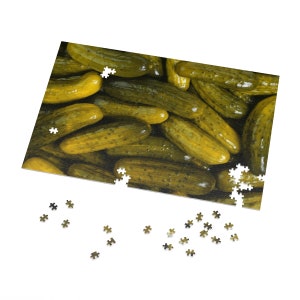 May include: A close-up image of a jigsaw puzzle with a picture of a pile of green dill pickles. The puzzle is partially assembled, with some pieces missing.
