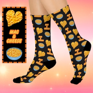 May include: A pair of black socks with a pattern of bowls of macaroni and cheese and hearts made of macaroni and cheese. The socks have the text "Mac & Cheese" repeated on them.