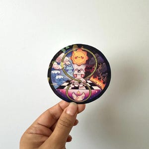 May include: A round sticker featuring a cartoon illustration of a cat-like creature in a yin-yang design. The design includes elements of sun, water, fire, and clouds, with a checkered floor and a crescent moon.