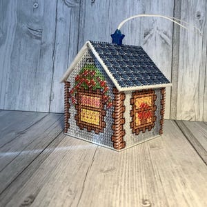 以下が含まれることがあります： 青いタイルの屋根と星型のトップが付いた、手作りのクロスステッチの家のオーナメント。窓には花柄とハートのデザインがあります。家は赤と緑の花柄のアクセントで飾られており、吊るすように設計されています。